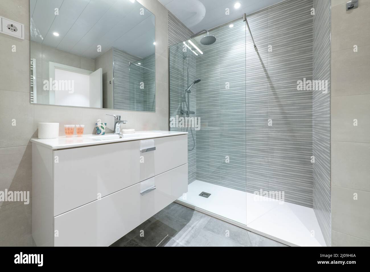 Salle de bains avec cabine de douche avec cloison en verre, lavabo mural en porcelaine blanche sur meuble en bois, miroir rectangulaire et carrelage en grès céramique Banque D'Images