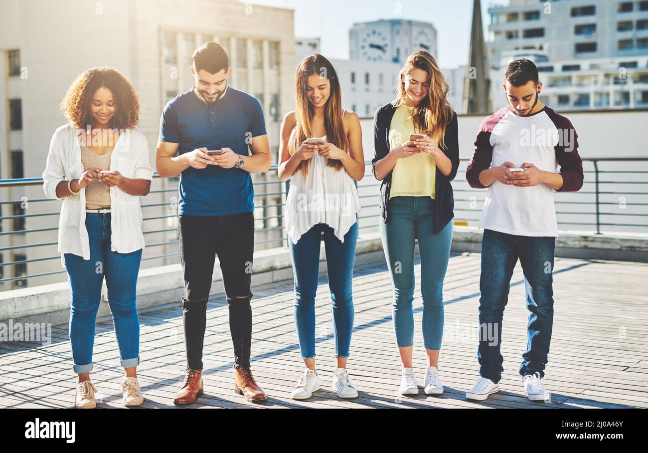 Connecté dans la ville. Prise de vue en longueur d'un groupe de jeunes qui envoient des SMS sur leur téléphone portable tout en étant debout à l'extérieur. Banque D'Images