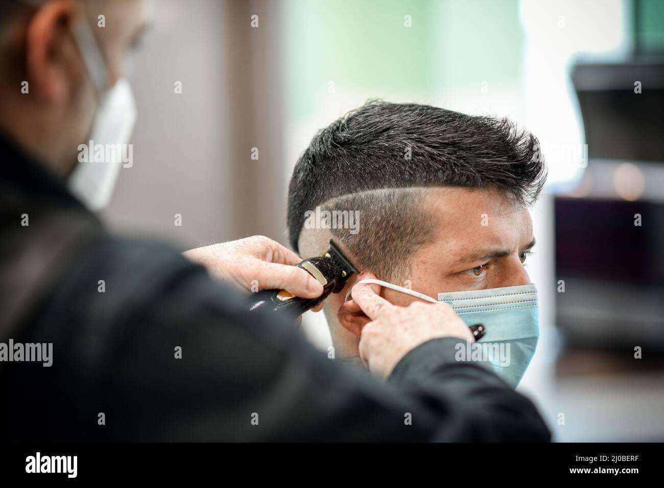 Crop barber dans le masque avec le coupe-cheveux faisant une coupe de cheveux à un client masculin dans le salon de coiffure pendant l'épidémie de COVID 19 Banque D'Images