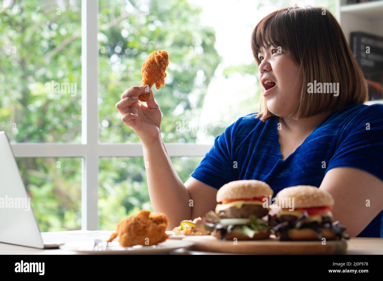 Une femme en surpoids affamée tenant le poulet frit, le hamburger sur une  assiette en bois et la pizza sur table, pendant le travail de la maison, de  prendre le problème de