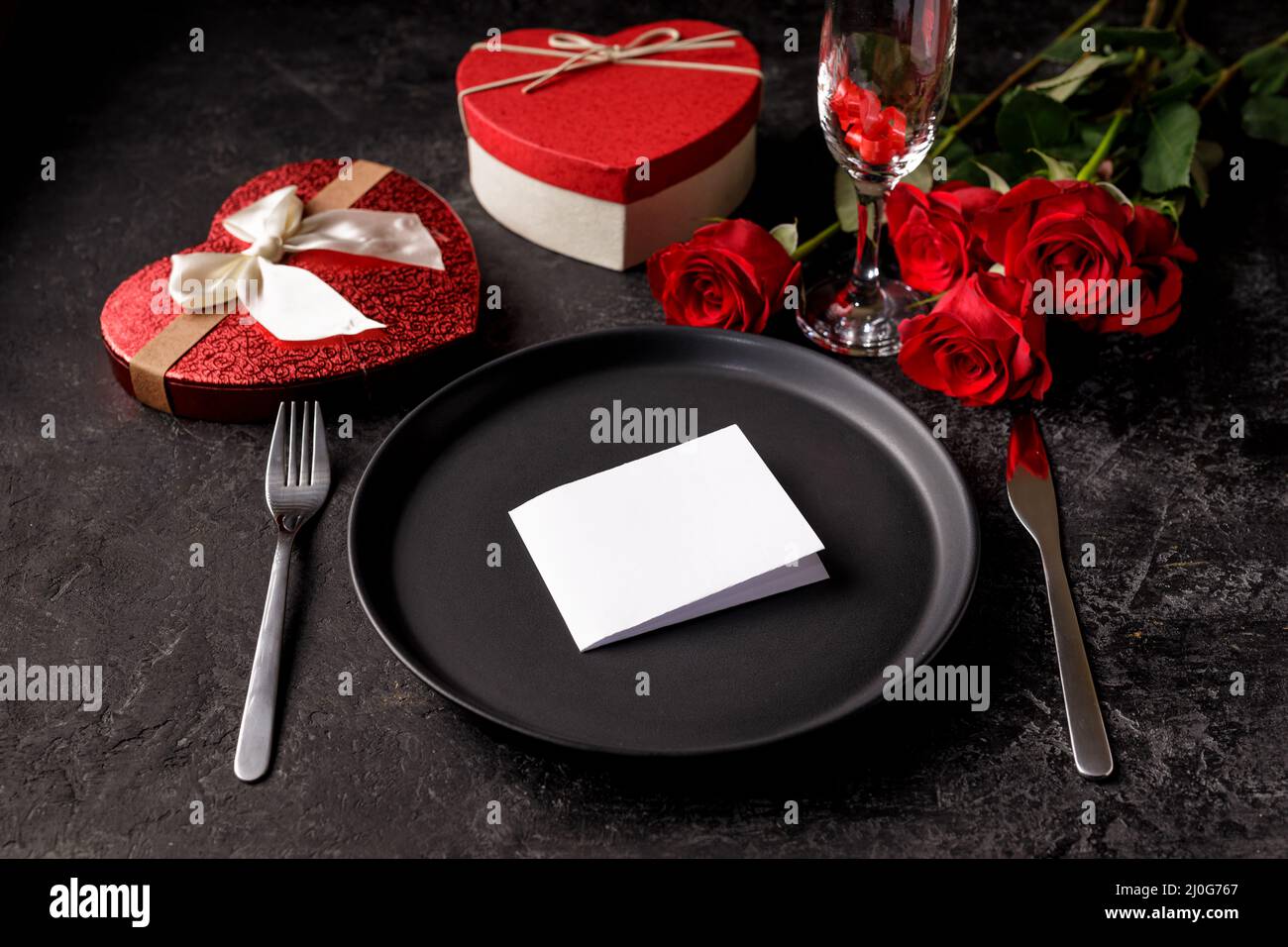 Dîner de fête de la Saint-Valentin ou du jour de naissance avec une assiette mate noire vide, une boîte cadeau en forme de foyer et des roses rouges Banque D'Images