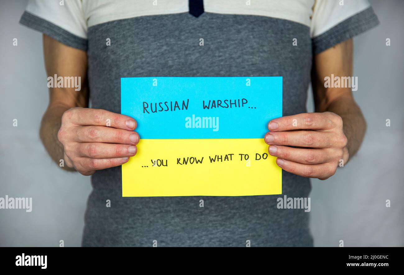 Un homme debout sur un gris, tient dans ses mains le fond d'un drapeau ukrainien en carton jaune-bleu avec l'inscription 'navire de guerre russe, vous savez quoi à Banque D'Images