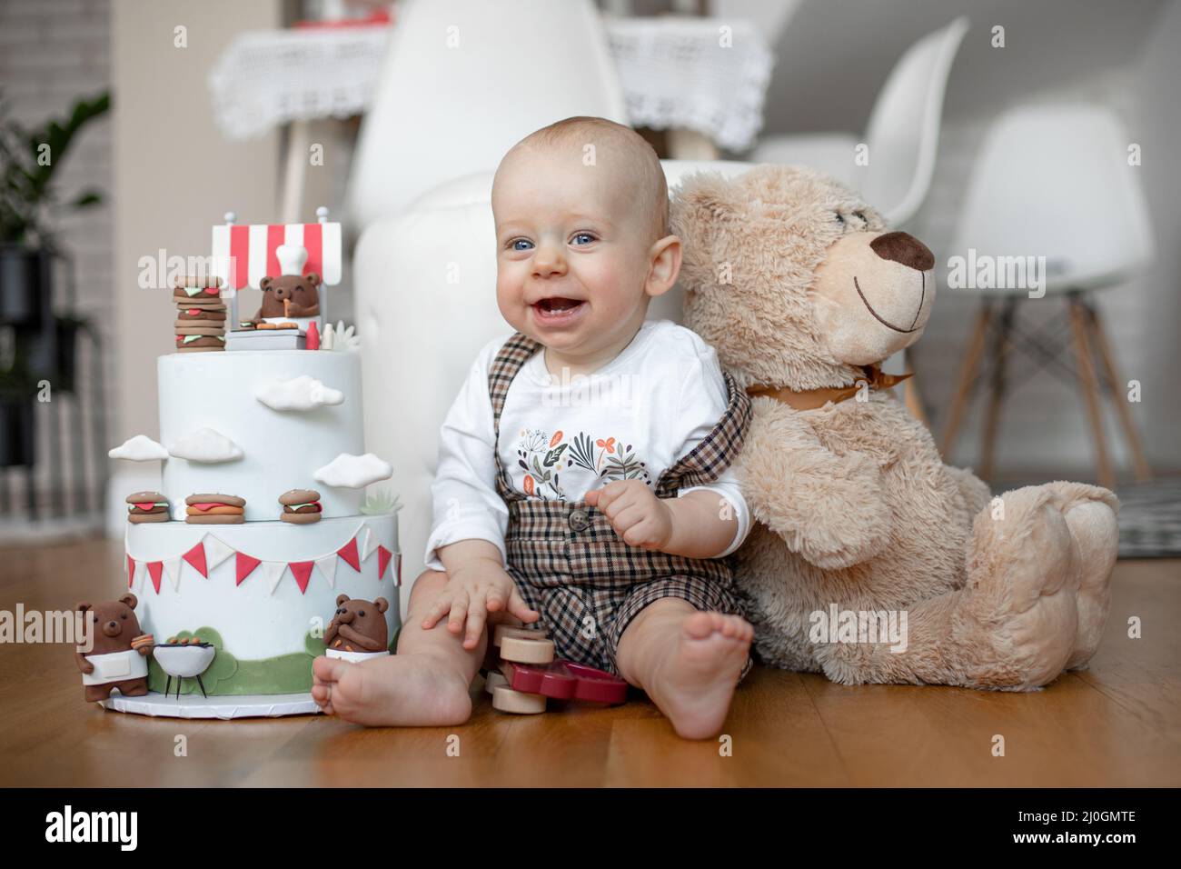 Petit garçon souriant en T-shirt blanc et à carreaux, assis sur le sol près d'un gâteau d'anniversaire à deux niveaux et d'un ours en peluche. Banque D'Images