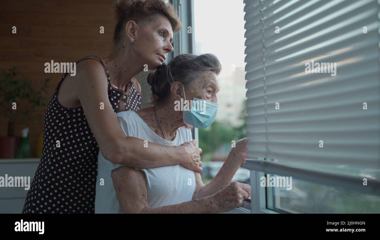 Femme solitaire âgée de 90 ans dans un masque médical et sa fille mature debout dans embrasse regardant la fenêtre dans la maison de soins infirmiers. Banque D'Images