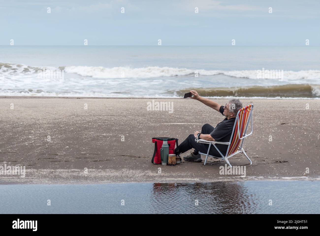 Homme mûr avec des cheveux gris et des lunettes assis sur une chaise de plage prenant un selfie. En arrière-plan la plage et les vagues de la mer. Banque D'Images