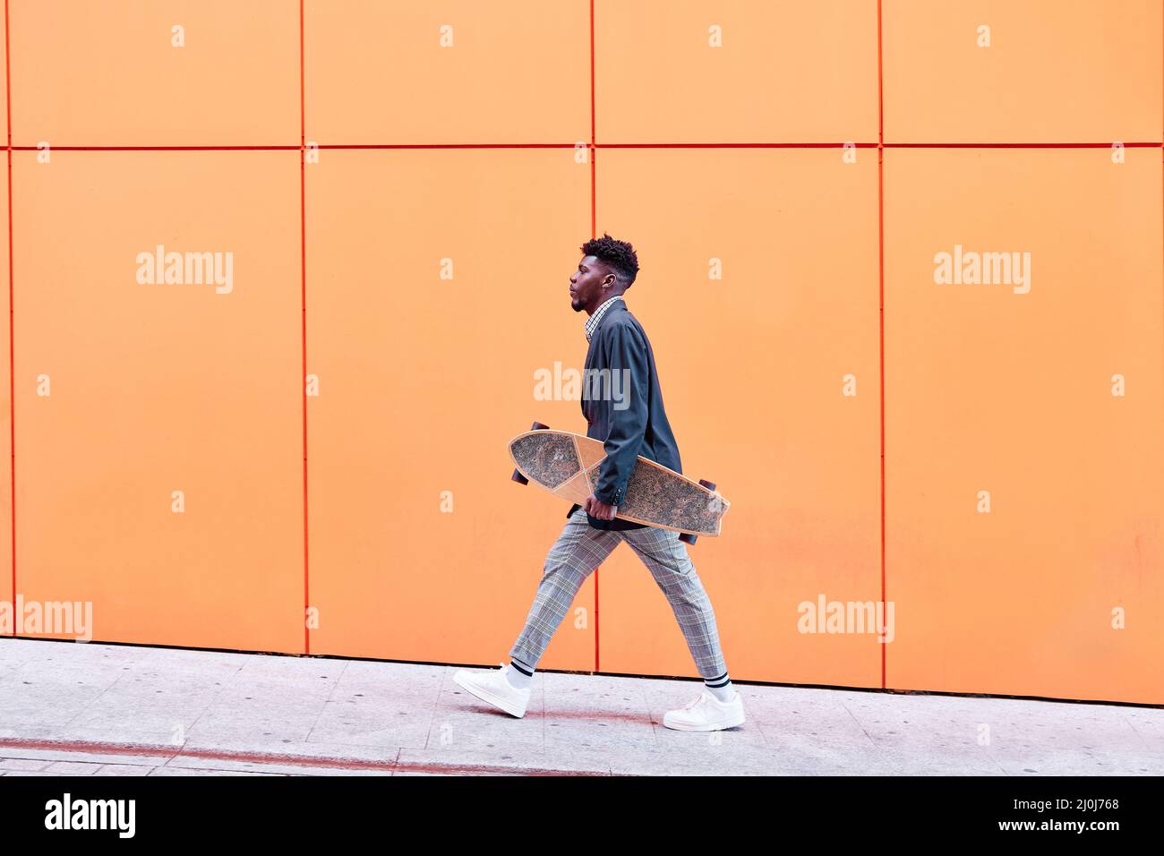 vue latérale d'un jeune homme avec blazer et planche à roulettes en bas de la rue sur fond orange. Banque D'Images