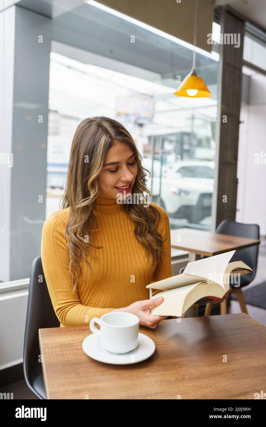 style de vie gai de jeune femme latine attrayante avec de longs cheveux blonds, appréciant la lecture d'un livre, à la table une tasse de café, passe-temps intellectuel Banque D'Images