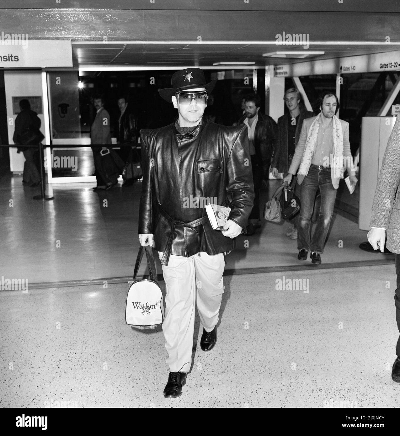 Elton John, le président et la star pop de Watford, a de bonnes raisons de se réjouir à bord d'un avion à Heathrow pour Antigua. Son côté a frappé Manchester United de la coupe FA à la veille, marquant le seul but du match. 3rd janvier 1982. Banque D'Images