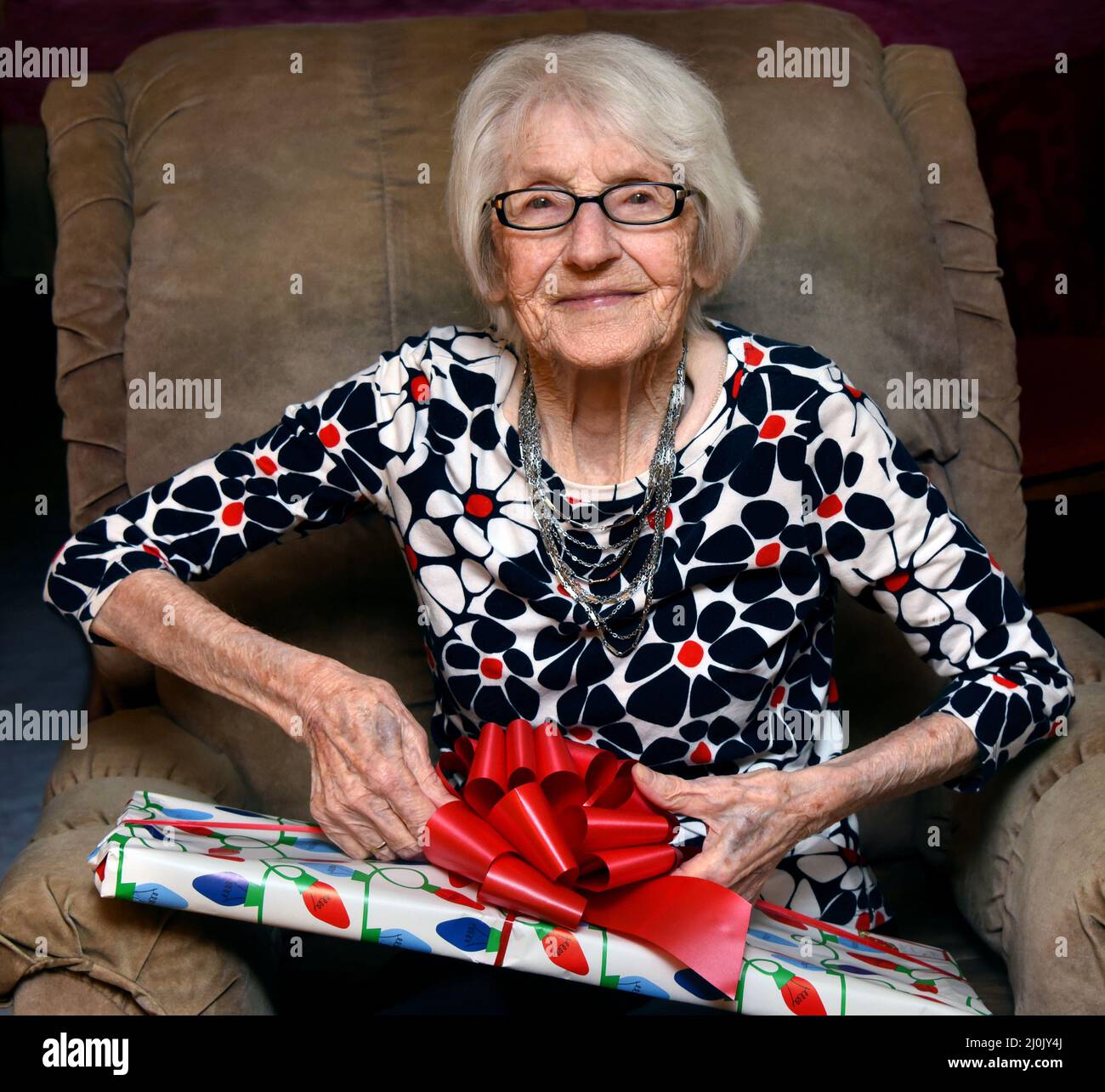 Une grand-mère de 100 ans s'assoit dans sa chaise préférée et ouvre un paquet coloré le matin de Noël.Elle regarde avec un sourire et scintillent en elle Banque D'Images