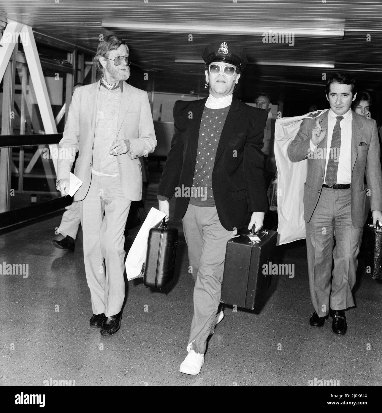 Le chanteur Elton John arrive à l'aéroport d'Heathrow après sa visite des États-Unis d'une durée de 10 semaines, portant un chapeau de policier américain. Sur la photo à droite se trouve son directeur John Reid. 21st août 1982. Banque D'Images