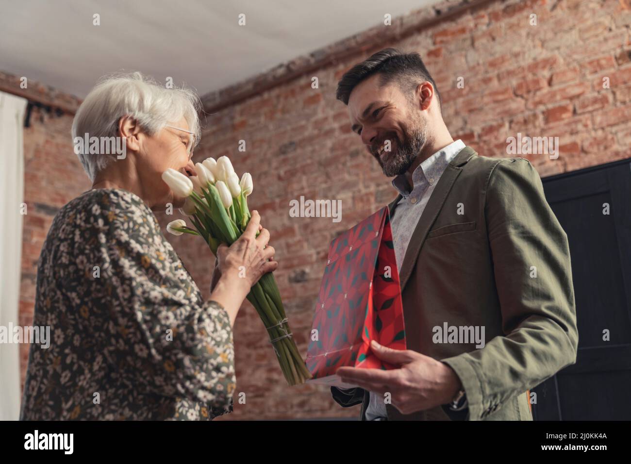 Femme caucasienne sénior aux cheveux gris ayant obtenu un bouquet de fleurs et un cadeau de son fils le jour de la fête des mères. Photo de haute qualité Banque D'Images