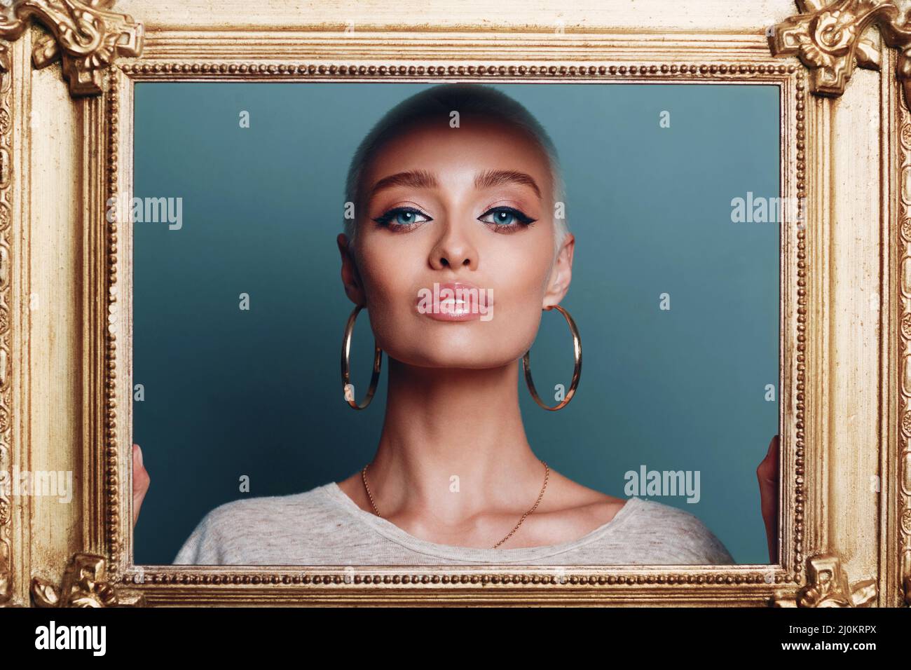 Jeune femme millénaire aux cheveux blonds courts tient le cadre doré dans les mains derrière son portrait du visage. Banque D'Images