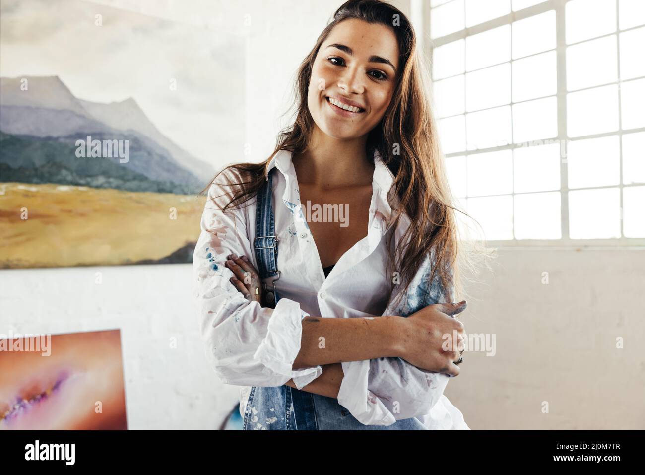 Joyeuse artiste souriant à la caméra tout en se tenant devant ses peintures. Heureuse peintre féminine debout avec ses bras croisés. Jeune créateur Banque D'Images