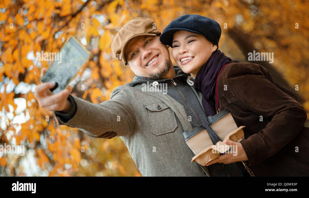 Couple souriant avec café à emporter fait Selfie dans le parc de la ville d'automne. Femme asiatique avec son Boyfriend va pour une promenade dans la FAL Banque D'Images