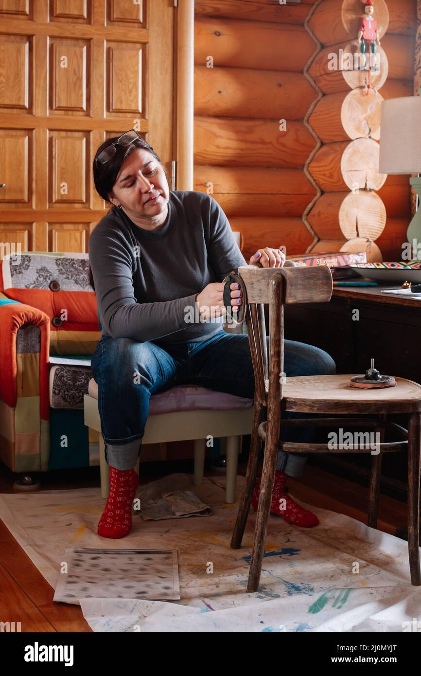 Portrait d'une femme d'âge moyen engagée à enlever la vieille couche de peinture de l'ancienne chaise avec un morceau de papier de verre dans l'atelier pour la rénovation des meubles Banque D'Images