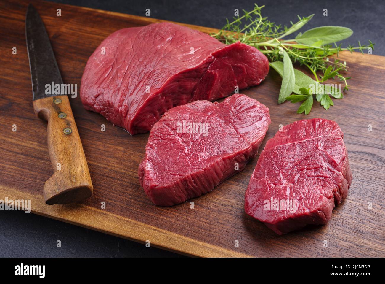 Morceau de steak de bosse de bœuf de bison sec et sec cru et tranches offertes avec des herbes en gros plan sur le plan de conception en bois Banque D'Images