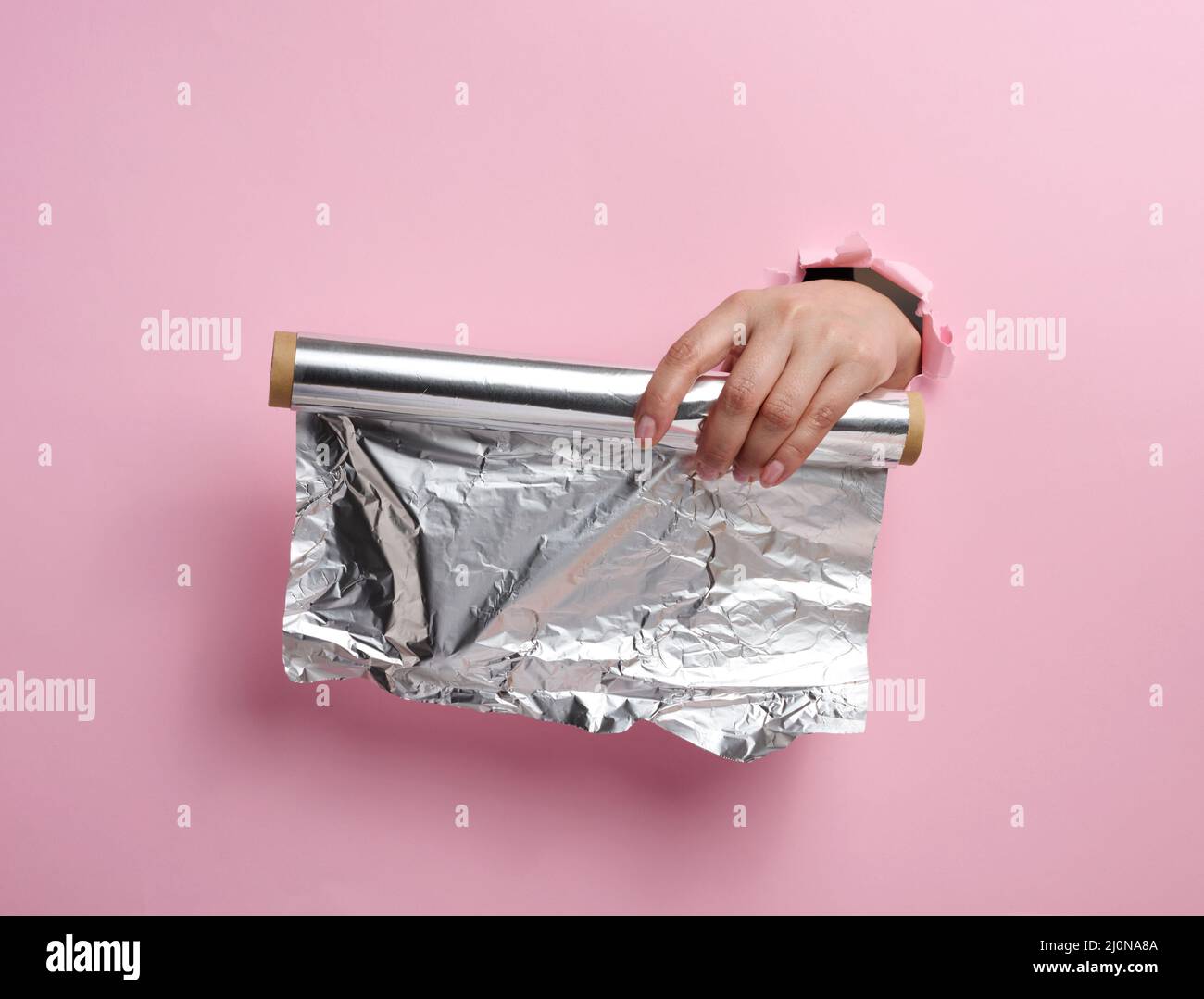 Main féminine tenant un rouleau de papier d'aluminium gris sur un fond rose, une partie du corps sortant d'un trou déchiré Banque D'Images