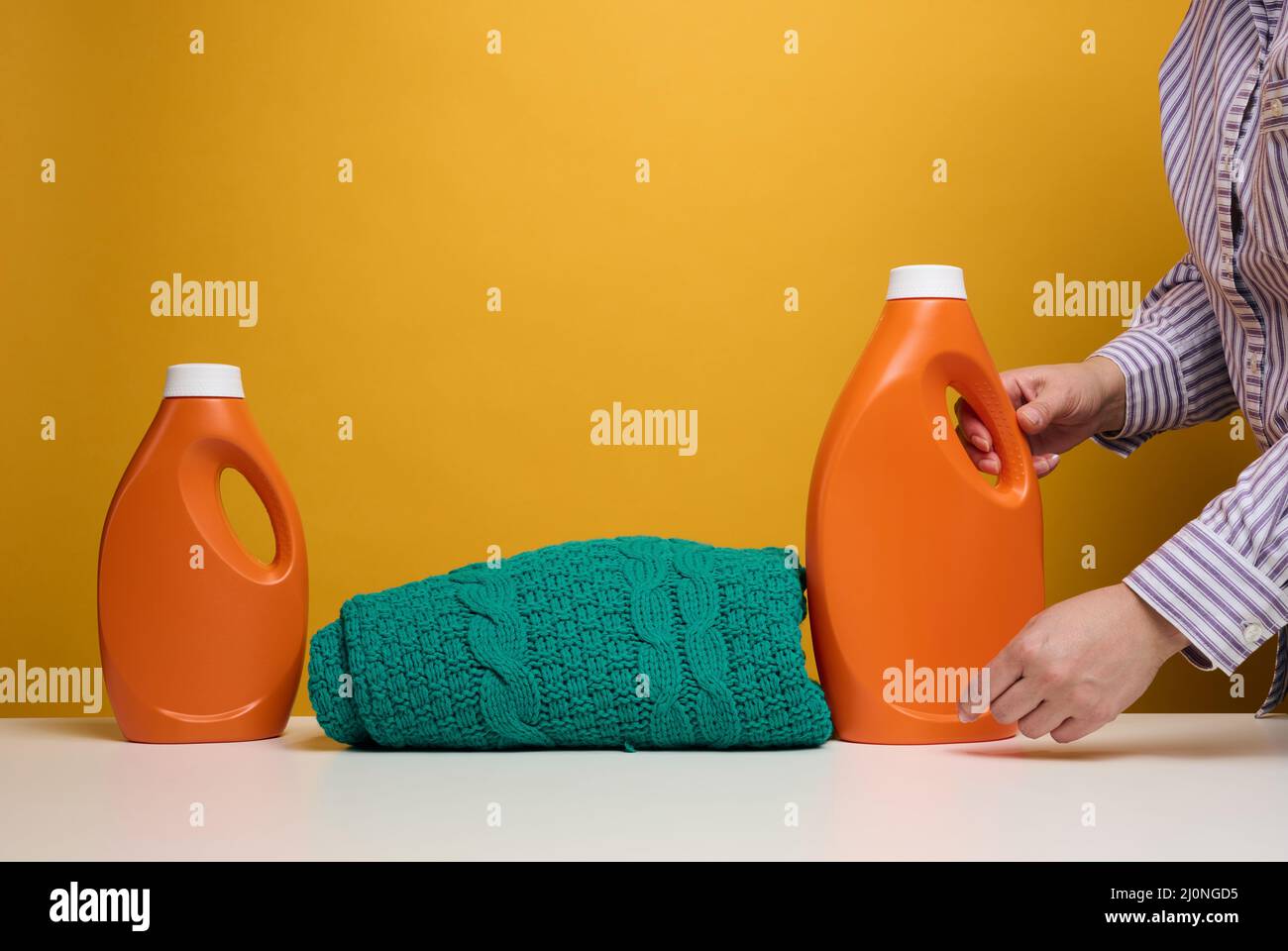 Vêtements pliés lavés et grandes bouteilles en plastique orange avec support de détergent liquide sur une table blanche, fond jaune. Routine Banque D'Images