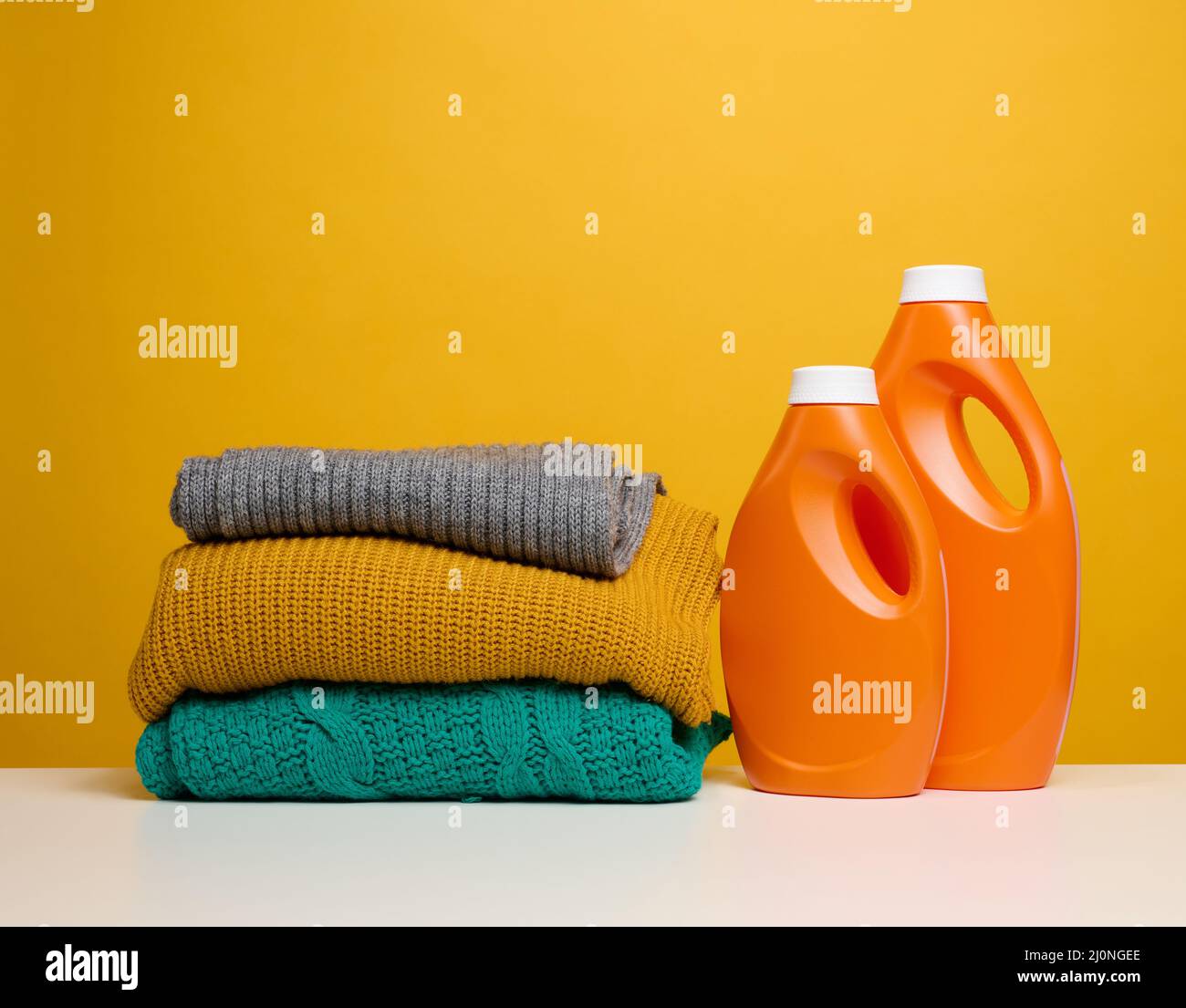 Pile de vêtements pliés lavés et grandes bouteilles en plastique orange avec détergent liquide sur une table blanche, fond jaune Banque D'Images