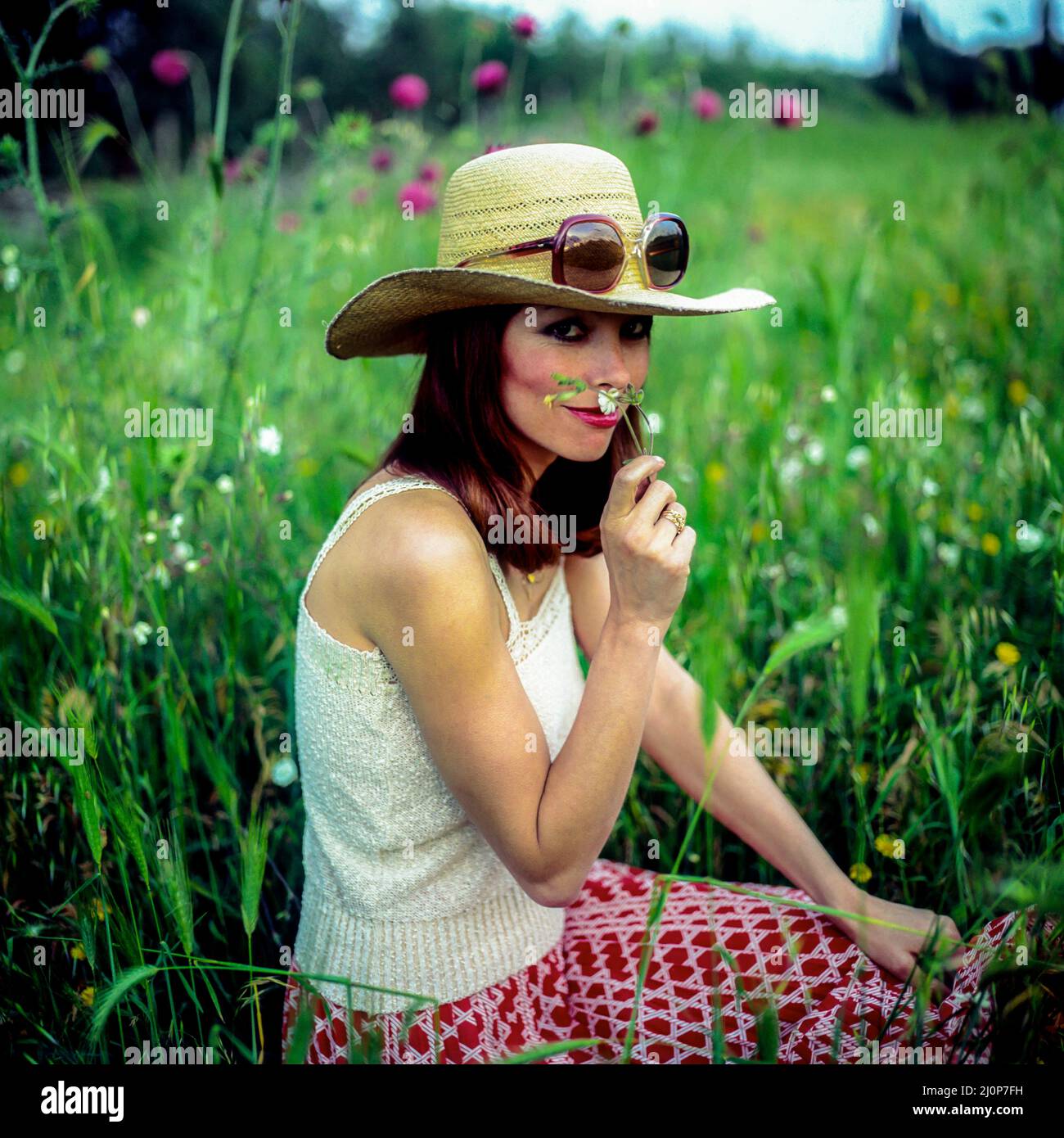 Vintage Italie 1970s, joli portrait de femme avec des lunettes de soleil sur chapeau de paille dans la prairie, odeur d'une fleur, Europe, Banque D'Images