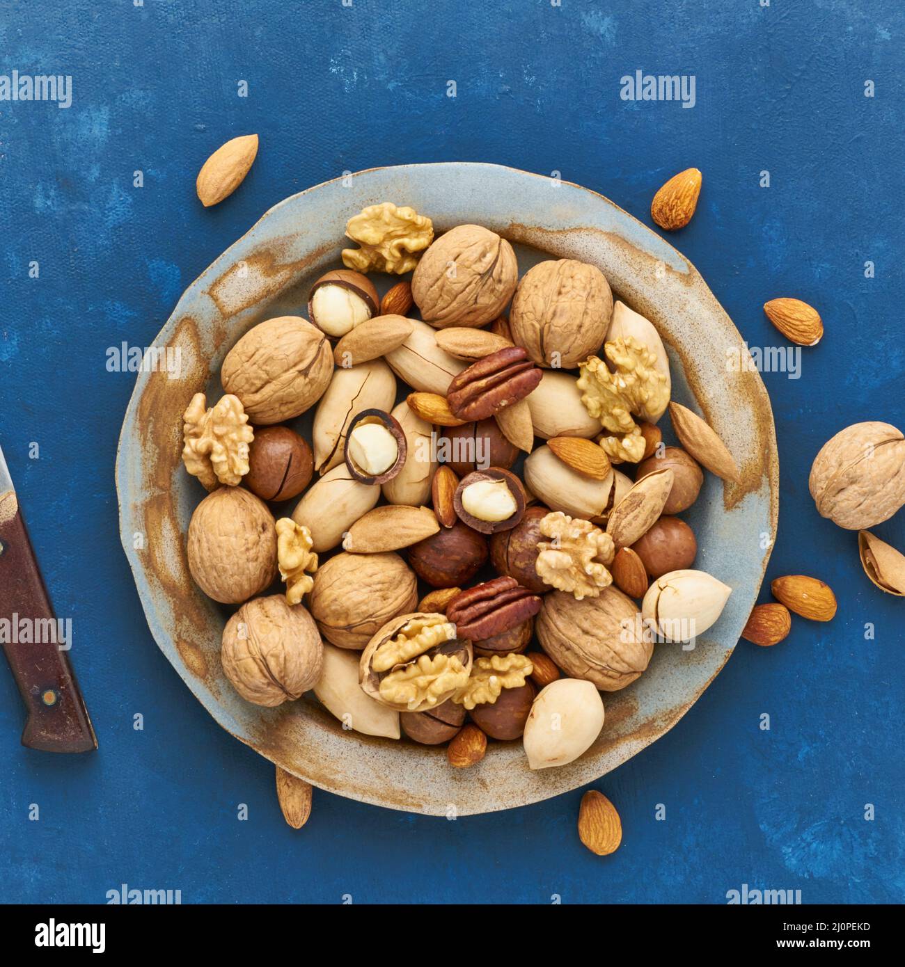 Bleu classique dans la nourriture. Mélange de noix sur l'assiette - noix, amandes, pacanes, macadamia et couteau pour ouvrir la coquille. Nourriture végétalienne saine. Banque D'Images