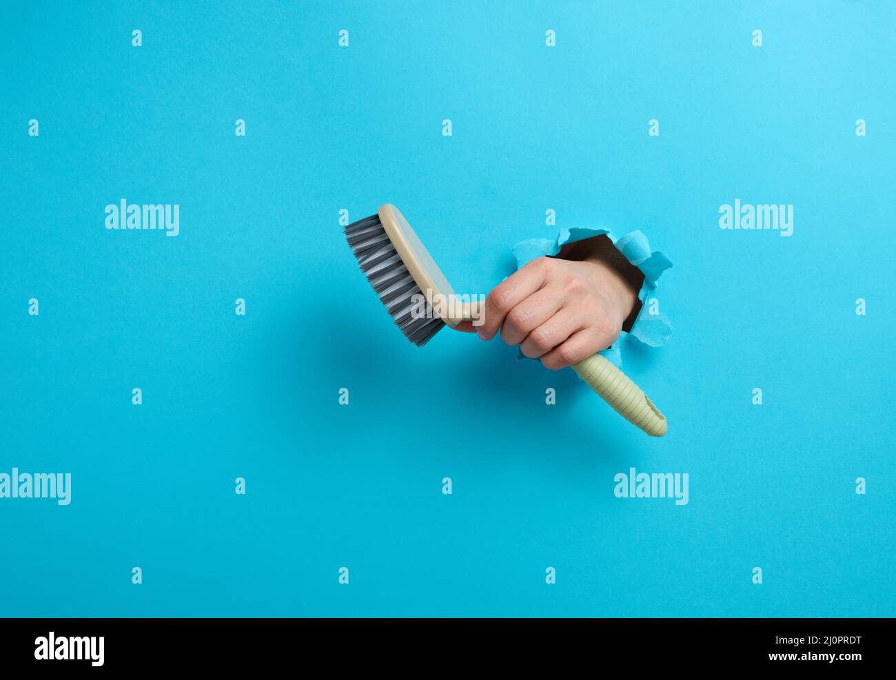 La main femelle tient une brosse en plastique grise sur fond bleu. Une partie du corps dépasse d'un trou dont les bords sont déchirés dans un papier Banque D'Images