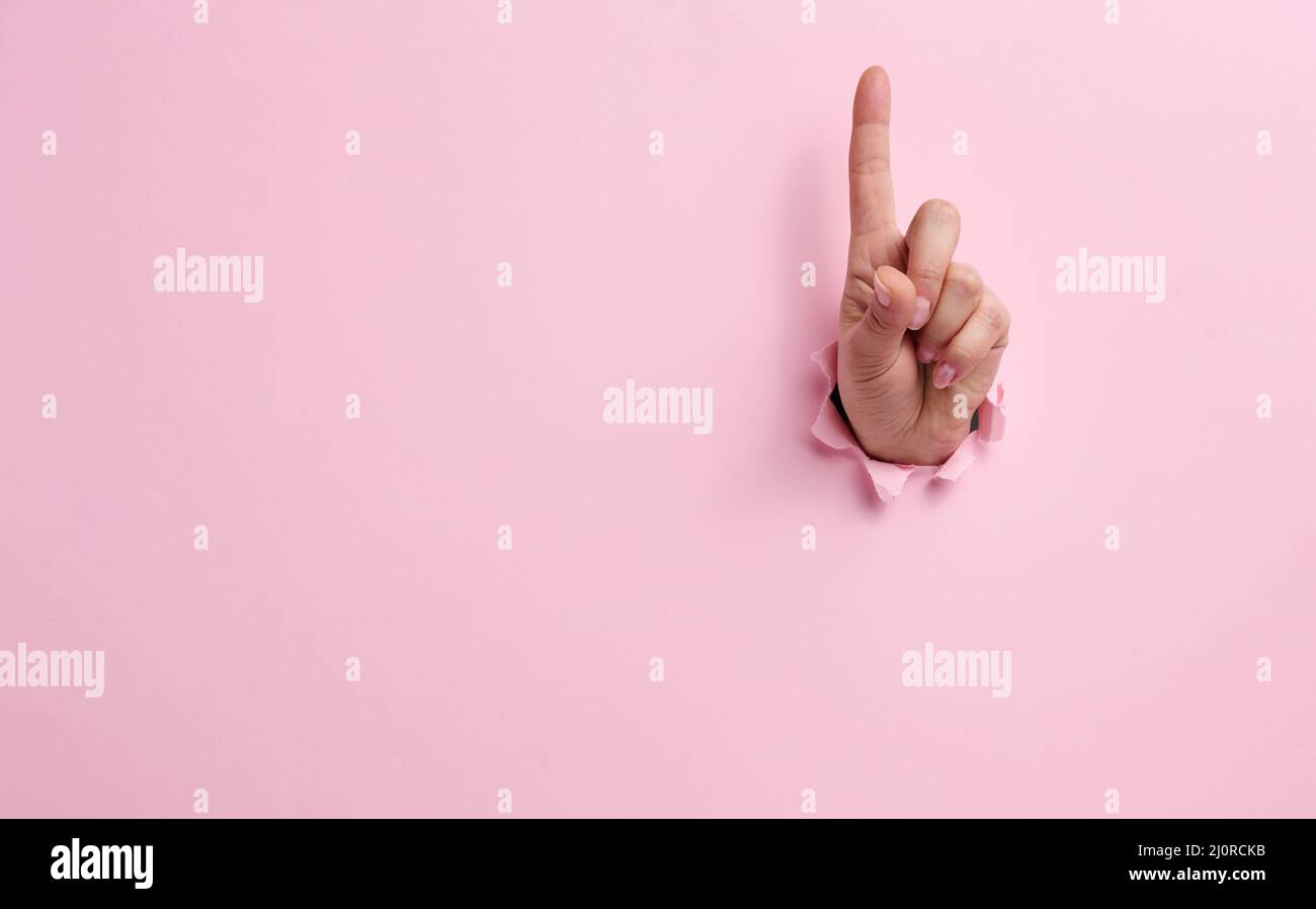 Main féminine qui dépasse d'un trou déchiré sur fond de papier rose, geste d'attention Banque D'Images