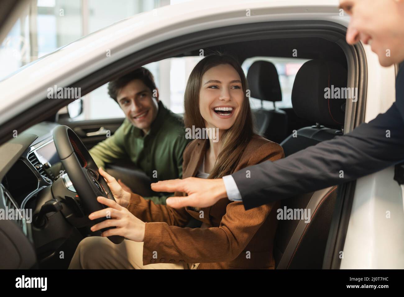 Un jeune couple heureux teste la conduite d'une nouvelle voiture, un vendeur l'aide à faire le meilleur choix chez la concession automobile Banque D'Images