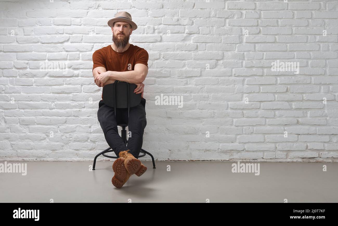 Homme barbu sérieux dans le chapeau assis devant le mur de brique blanche..bras croisés. Photo pleine longueur avec espace de copie. Banque D'Images