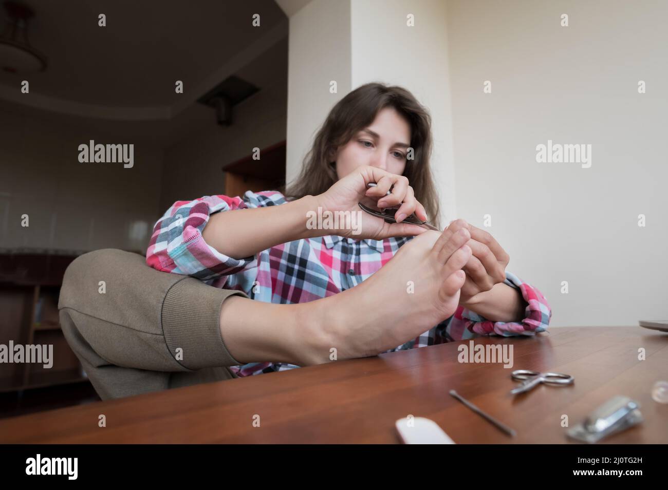 Belle jeune femme caucasienne dans une chemise assise dans la cuisine se faisant une pédicure. Soins à domicile en intérieur Banque D'Images