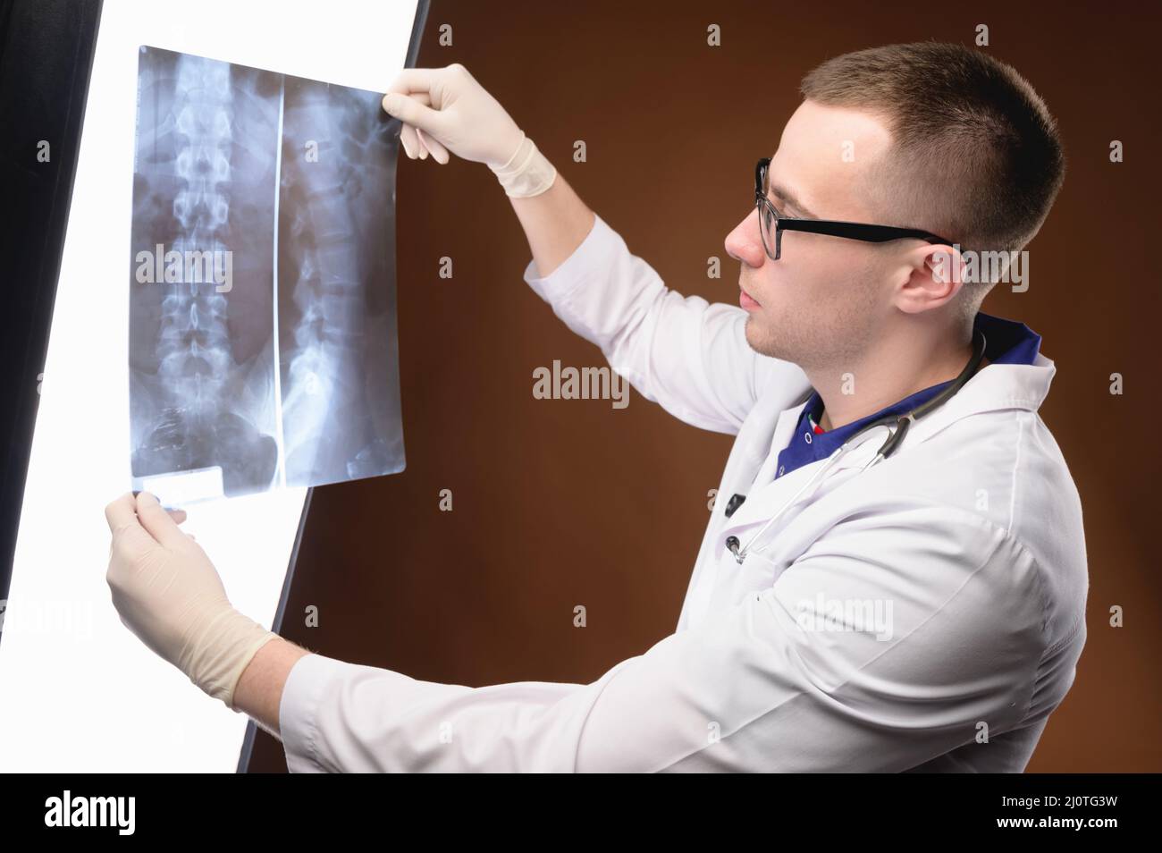 Le jeune médecin radiologue en lunettes et en vêtements médicaux regarde l'image de rayons X de la lumière de la colonne vertébrale sur fond marron. Banque D'Images