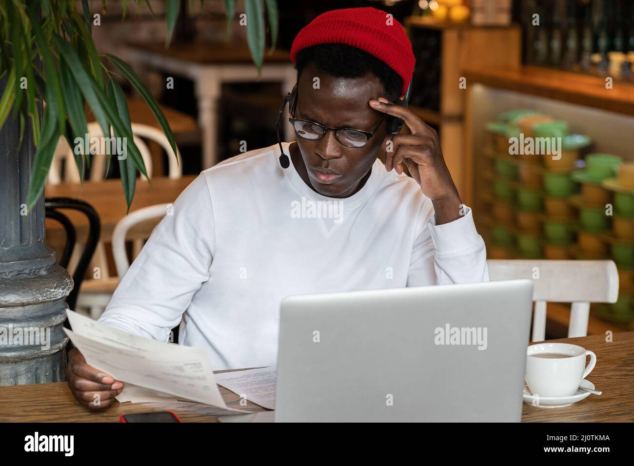 Pensive Afro-américain hipster homme dans des lunettes distance de travail sur ordinateur portable avec des documents dans le café Banque D'Images