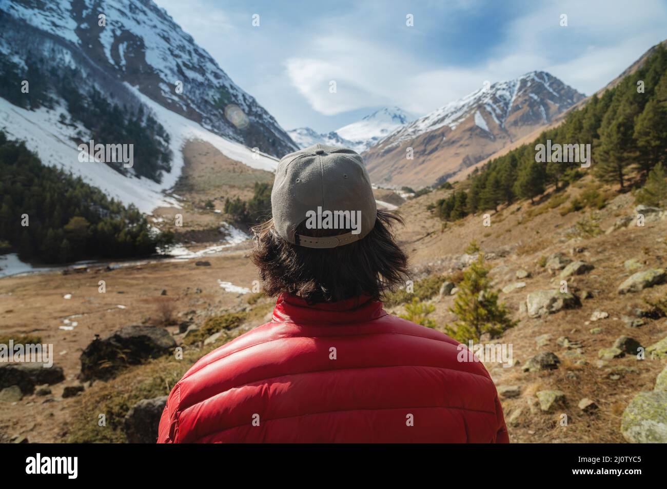 Un jeune homme aux cheveux longs, vêtu d'une veste en duvet rouge et d'une casquette grise en lunettes de soleil, se tient avec son dos à l'appareil photo et regarde une mounta Banque D'Images