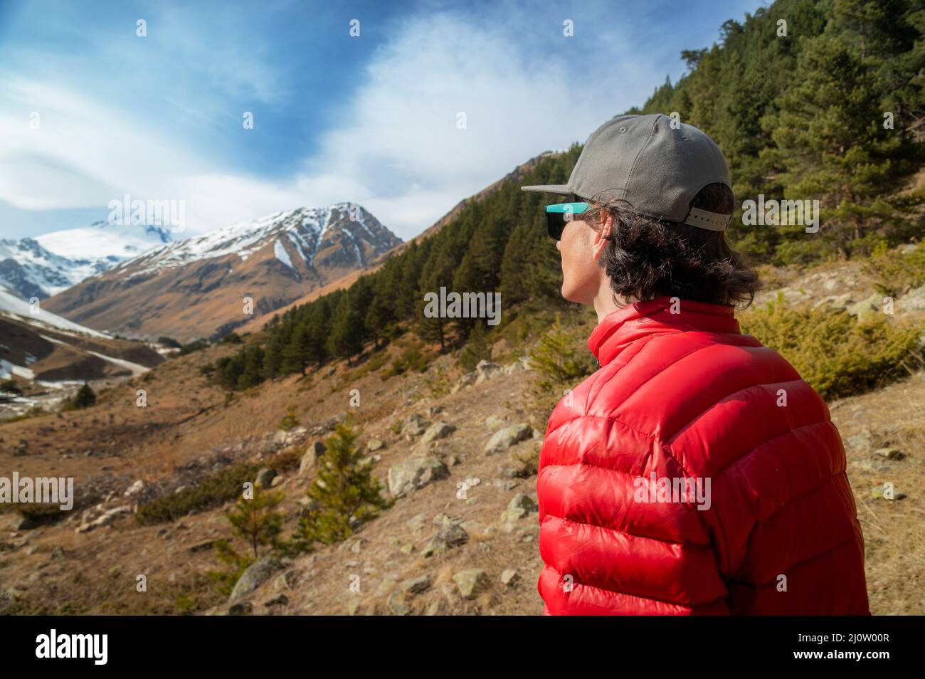 Un jeune homme aux cheveux longs, vêtu d'une veste en duvet rouge et d'une casquette grise en lunettes de soleil, se tient avec son dos à l'appareil photo et regarde une mounta Banque D'Images