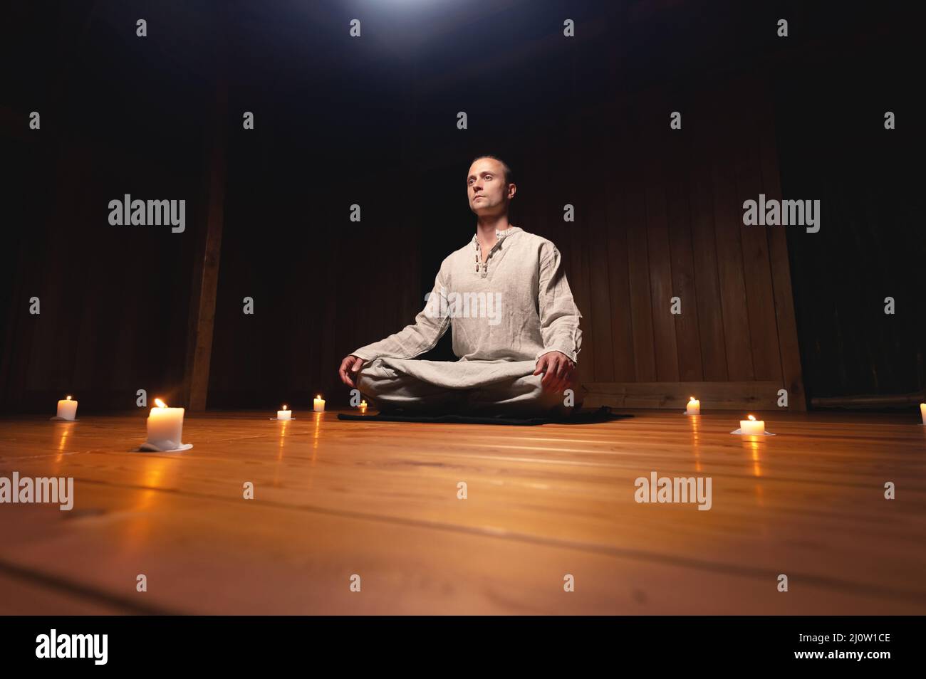 Un portrait d'un homme caucasien attrayant est assis dans des robes de coton dans une pose de lotus dans une salle d'entraînement en bois sombre entourée de céd Banque D'Images