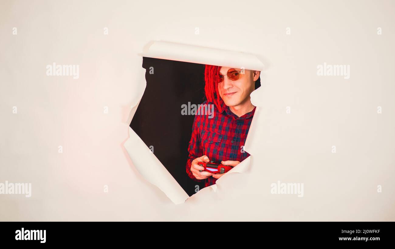 Impressionnant hipster en lunettes avec rouge dreadlocks joueur jouant des jeux d'ordinateur à travers un trou dans le mur blanc dans le studio Banque D'Images
