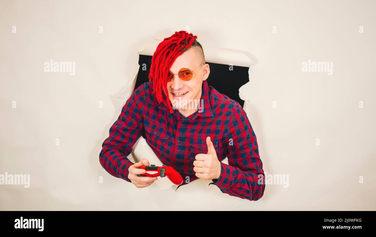 Impressionnant hipster en lunettes avec rouge dreadlocks joueur jouant des jeux d'ordinateur à travers un trou dans le mur blanc dans le studio Banque D'Images