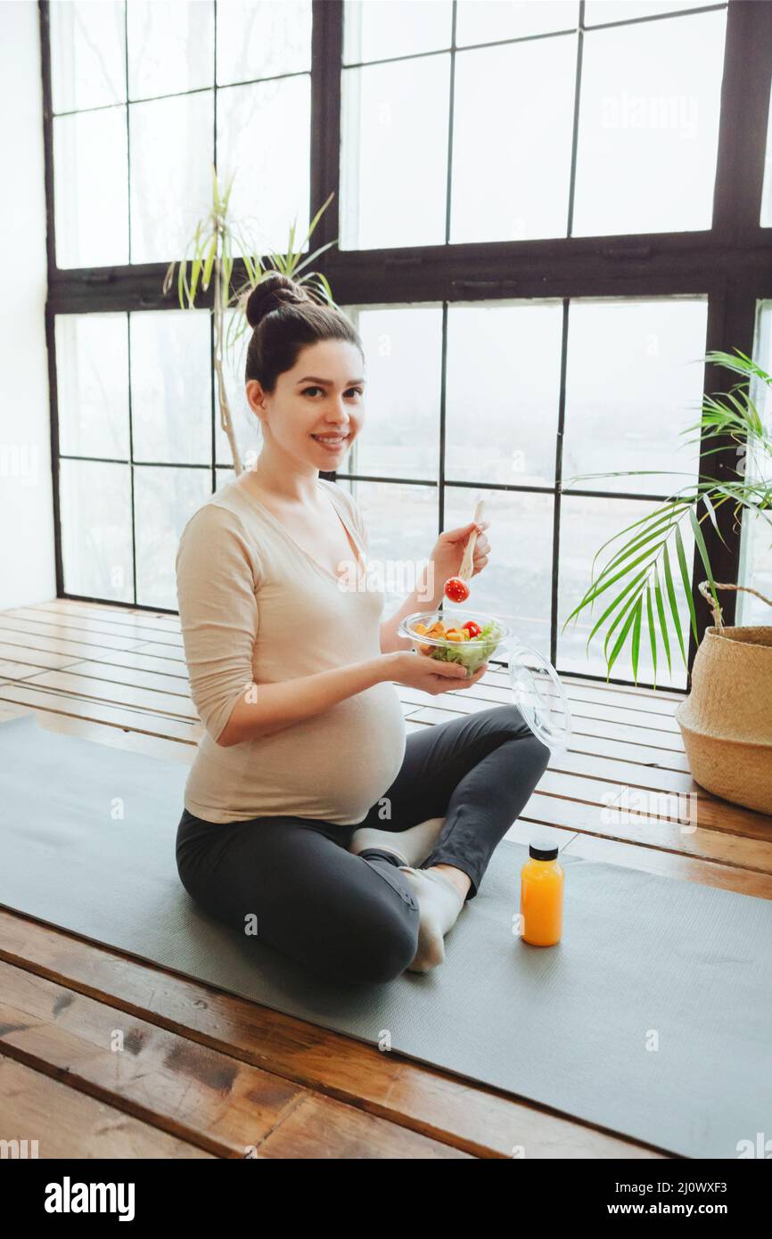 Un mode de vie sain et une alimentation saine pendant la grossesse. Une jeune femme enceinte mange de la salade sur un tapis de yoga Banque D'Images