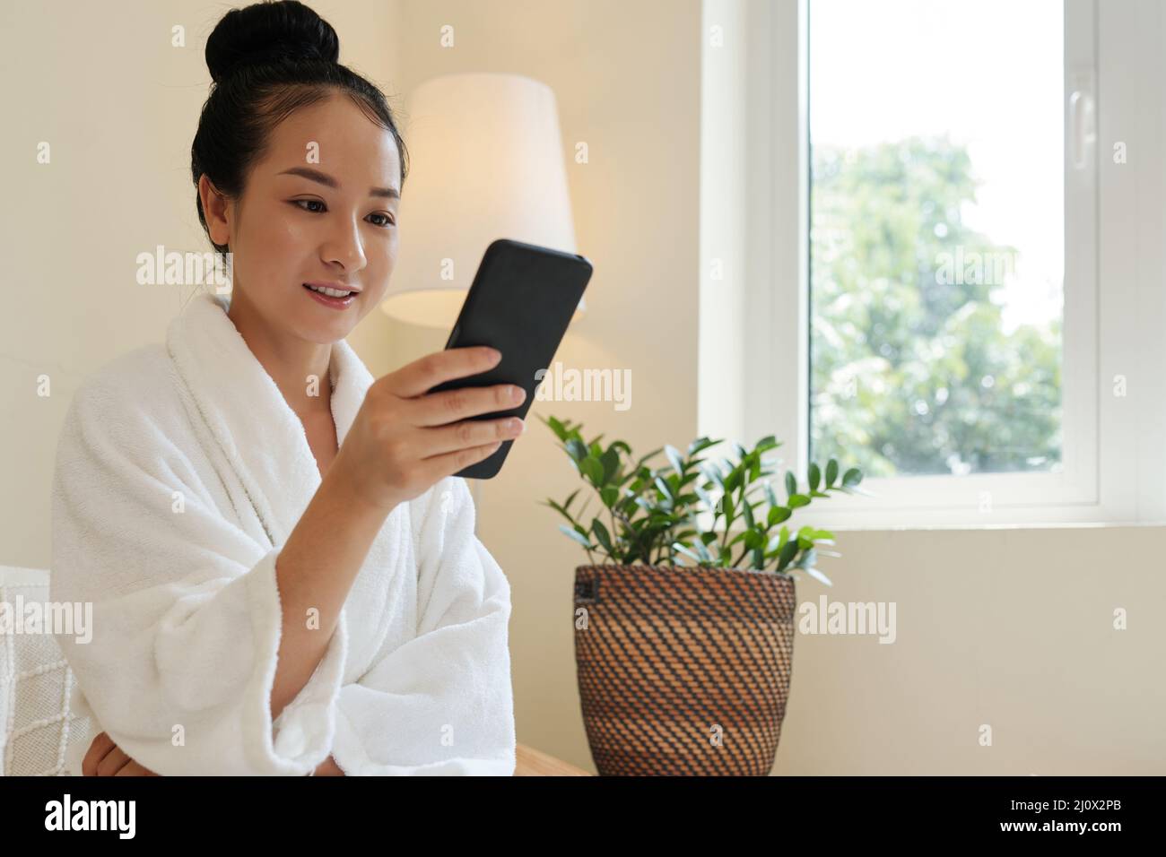 Jeune femme souriante en peignoir lisant des messages texte et des notifications sur smartphone Banque D'Images