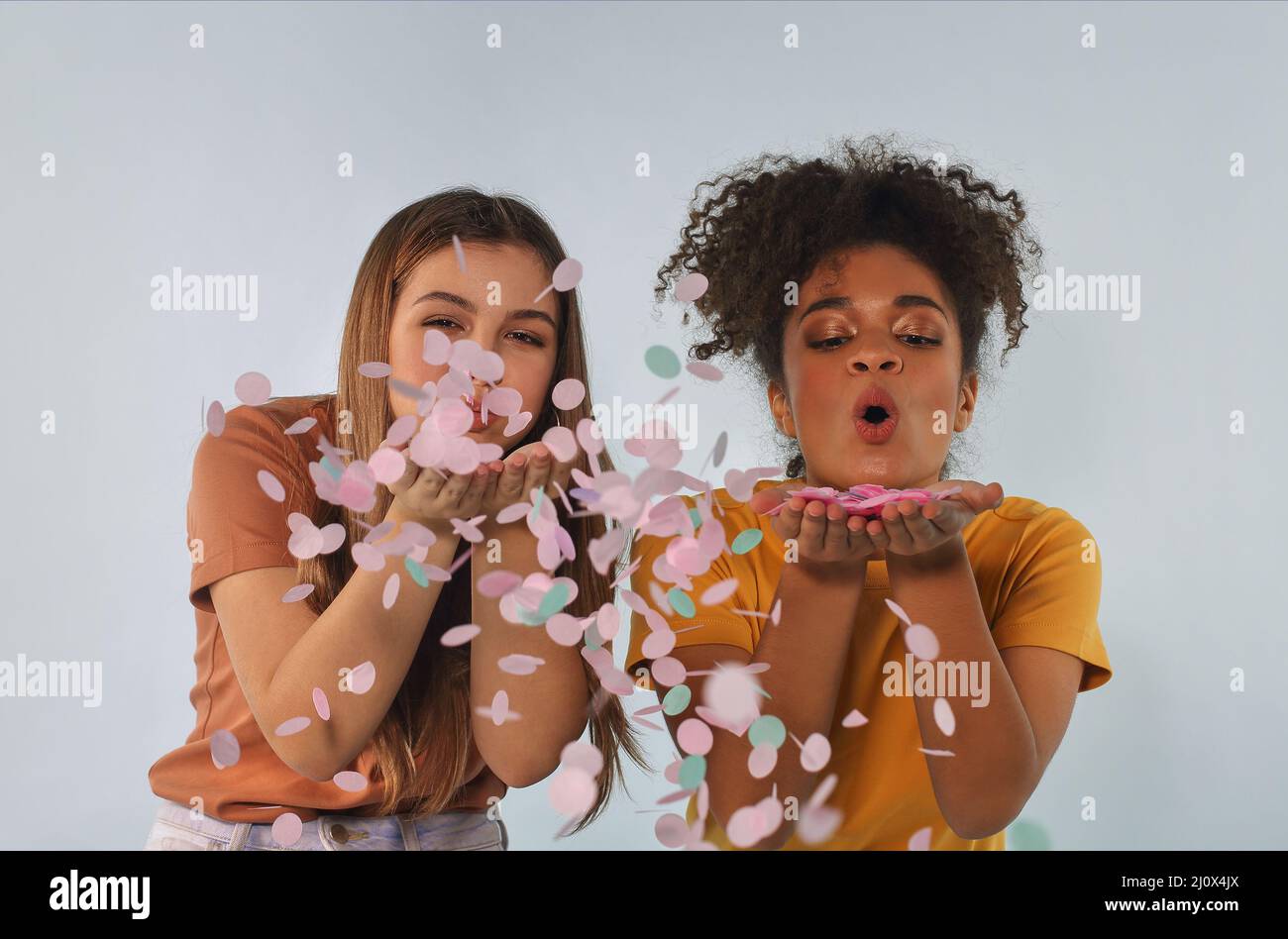 Deux amies multiraciales joyeuses qui soufflent des confettis à l'appareil photo et s'amusent, appréciant la fête d'anniversaire Banque D'Images