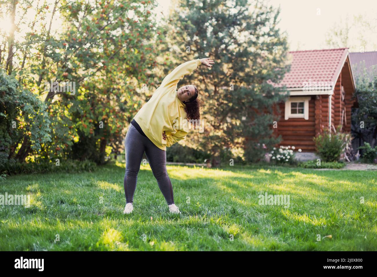 Femme étirant les bras sur l'herbe verte faisant des exercices physiques dans les vêtements de sport regardant la caméra sur l'arrière-cour avec maison de campagne et de grands arbres dedans Banque D'Images