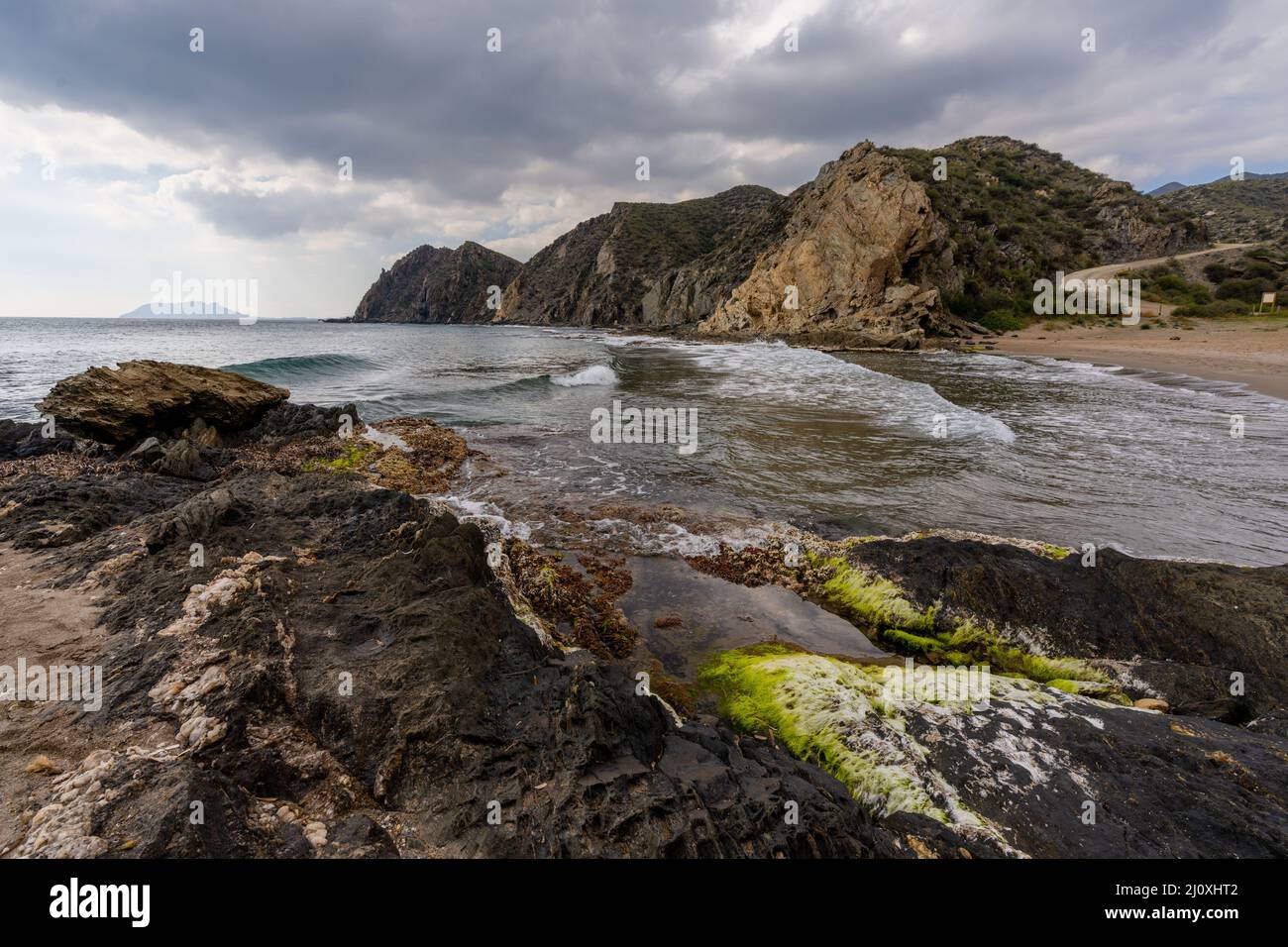 Une plage de sable isolée sur une côte montagneuse sauvage avec des rochers colorés et des algues au premier plan Banque D'Images