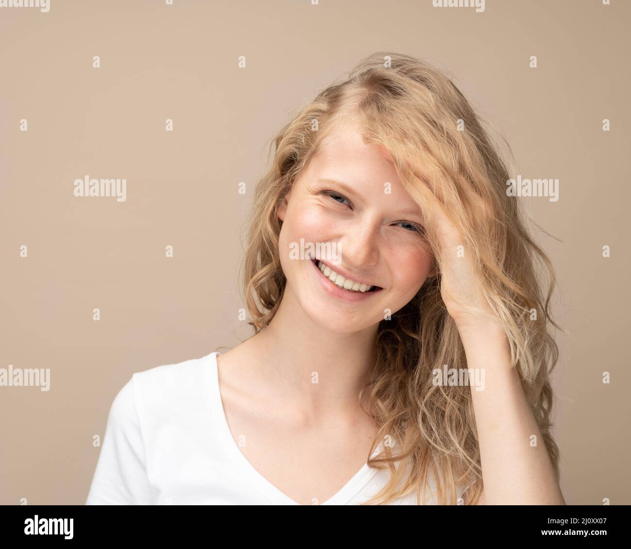 Belle jeune fille riant. Jolie blonde avec des cheveux bouclés dans un t-shirt blanc contre un mur beige souriant Banque D'Images