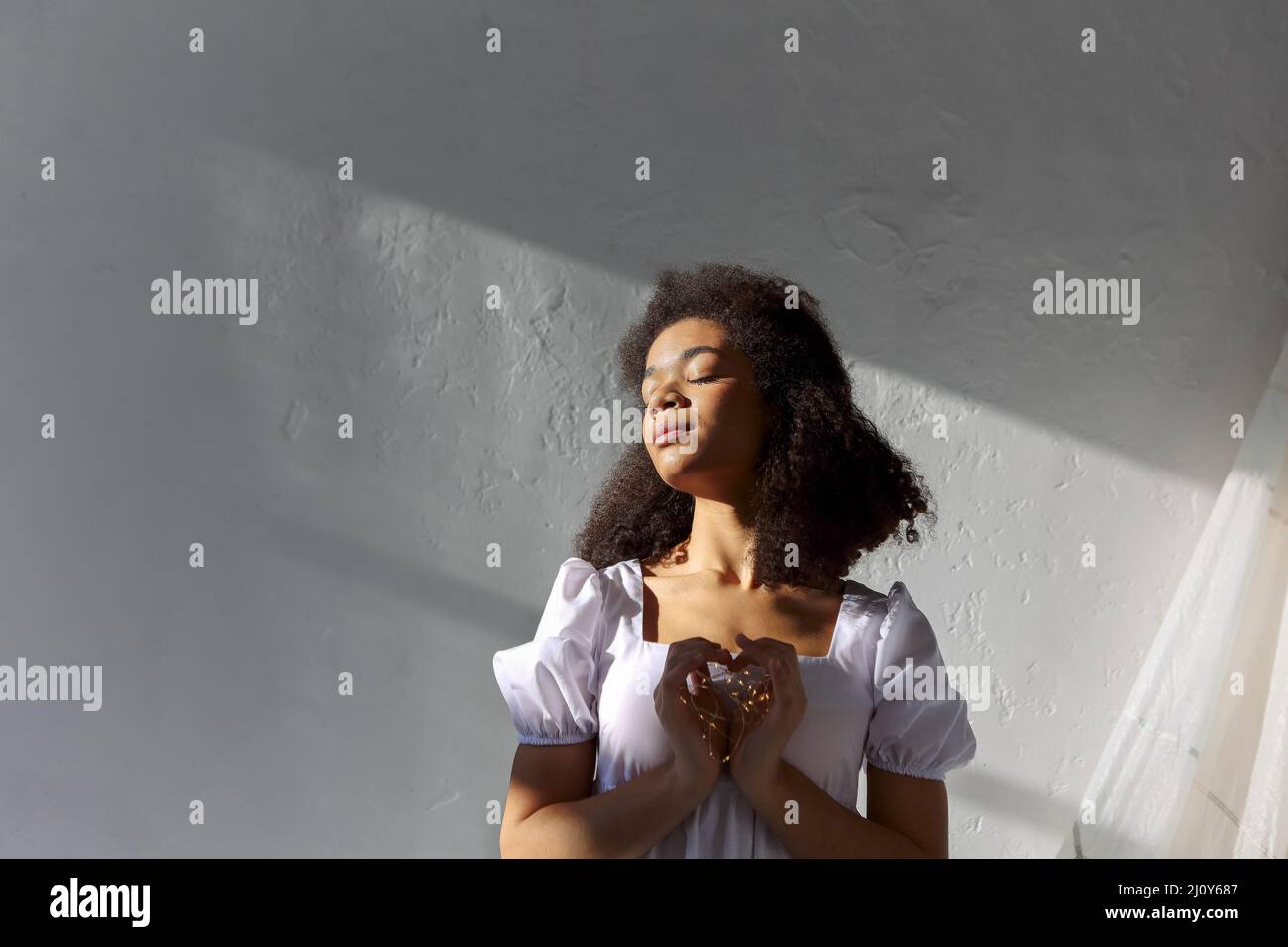 Jeune paisible reconnaissant afro-américaine reconnaissante femme avec les yeux fermés dans la robe blanche faisant signe de coeur, LED lumières dans les mains o Banque D'Images