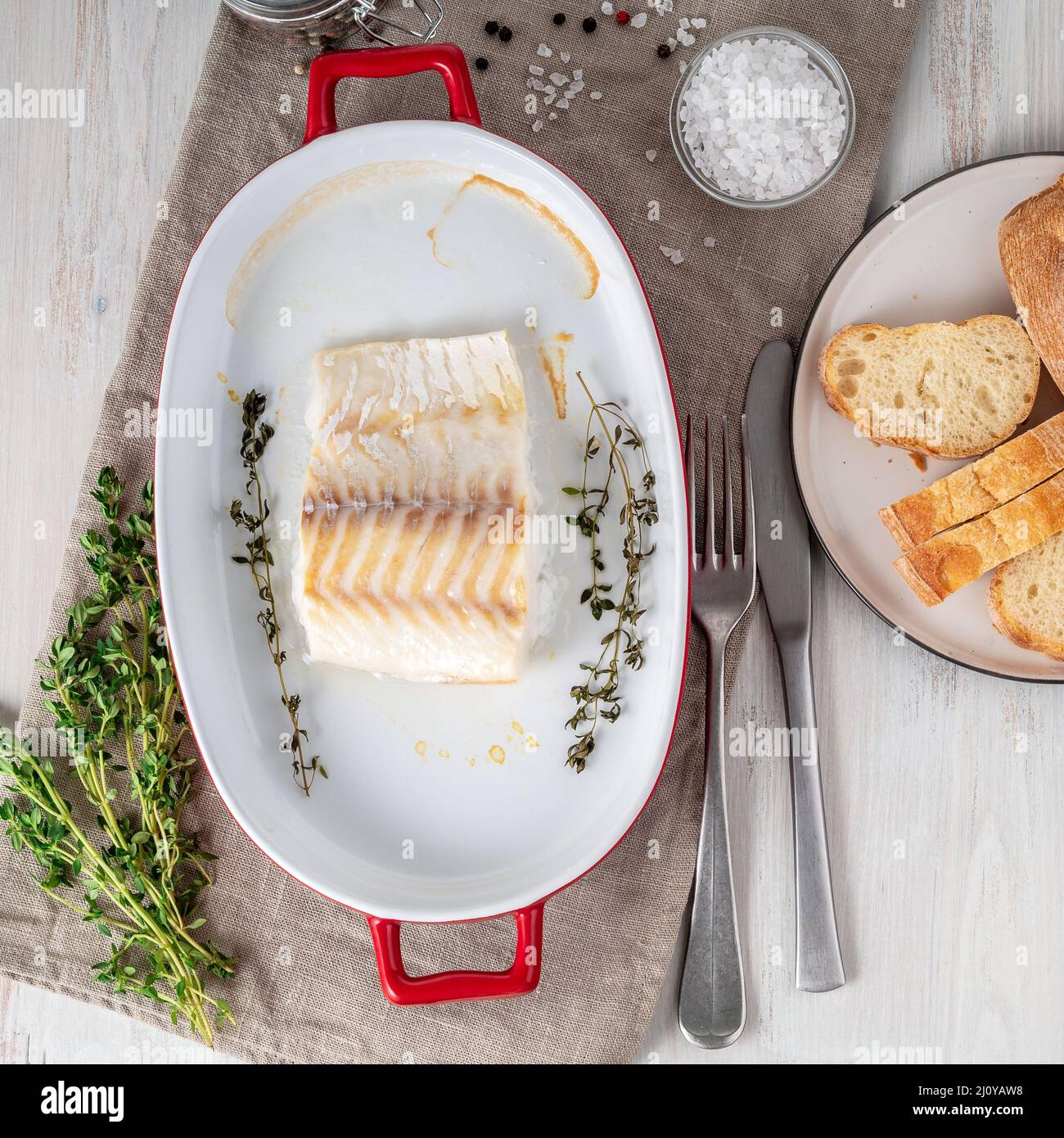 Filet de poisson de morue, fraîchement cuit au four avec du thym dans un plat en porcelaine pour la cuisson, pain sur une assiette, sel et poivre, bois blanc Banque D'Images