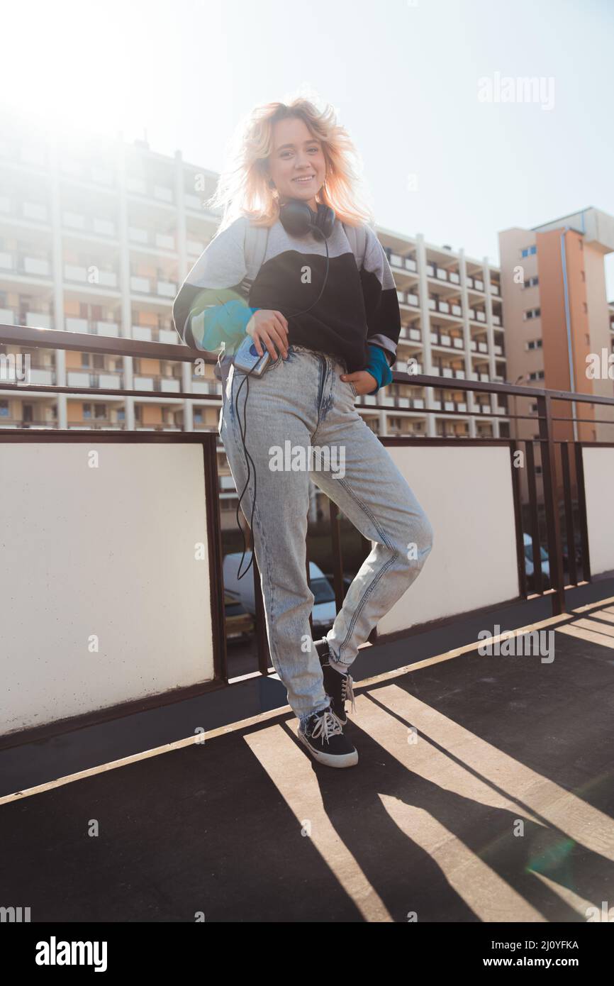 90s vibes. Jeune fille européenne avec de grands écouteurs noirs sur le cou, regardant l'appareil photo et souriant. Prise de vue verticale en extérieur. Photo de haute qualité Banque D'Images