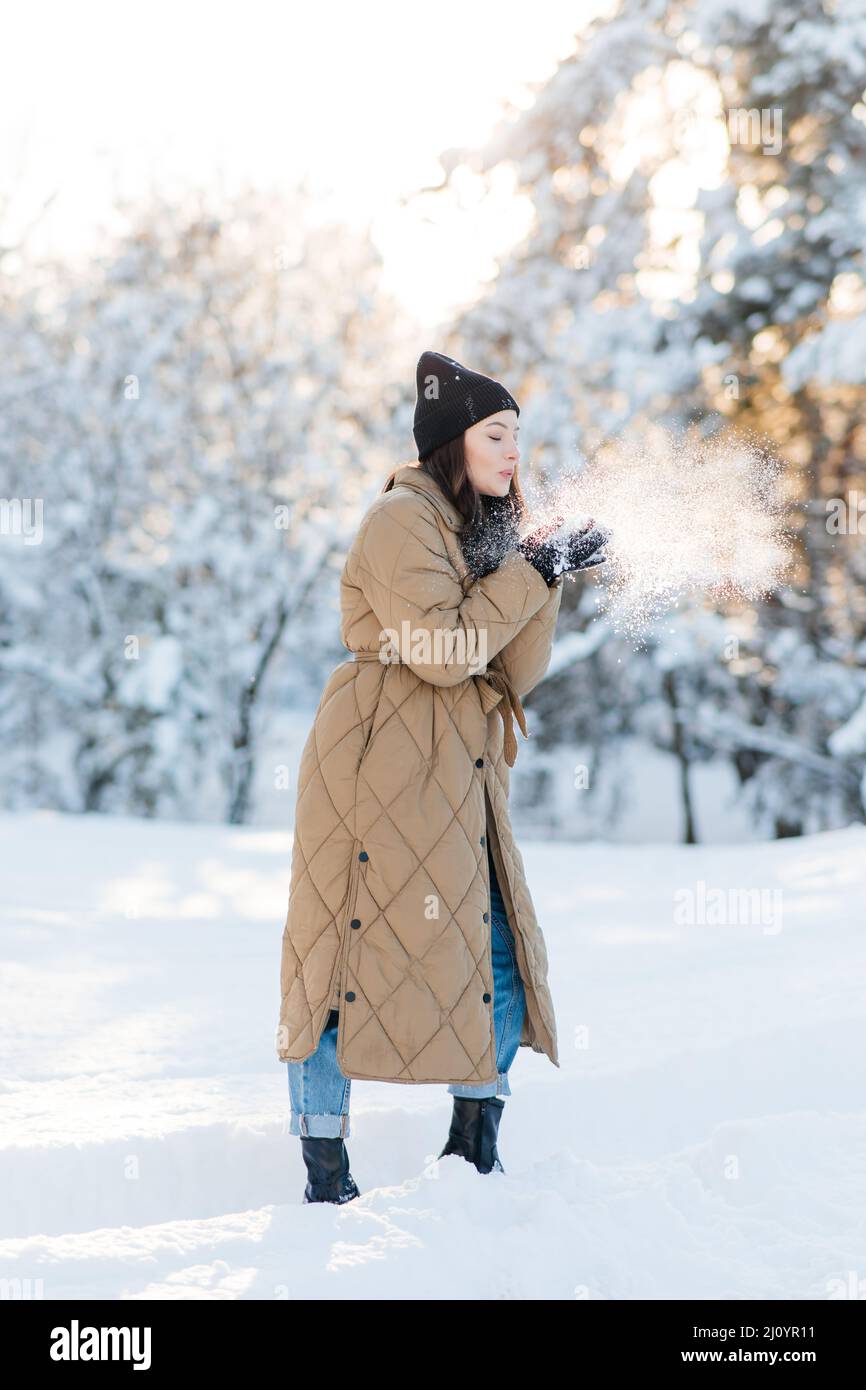 Bonne jeune femme souriante marche et souffle sur la neige dans ses mains dans la forêt parmi les pins couverts de neige dans la journée ensoleillée d'hiver. Banque D'Images