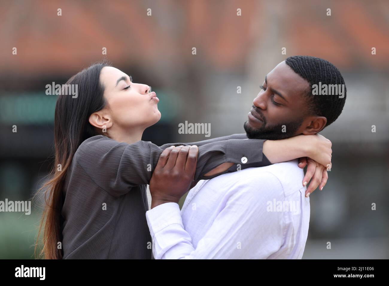 Vue latérale portrait d'une femme obsédée essayant d'embrasser un homme à la peau noire qui la rejette dans la rue Banque D'Images