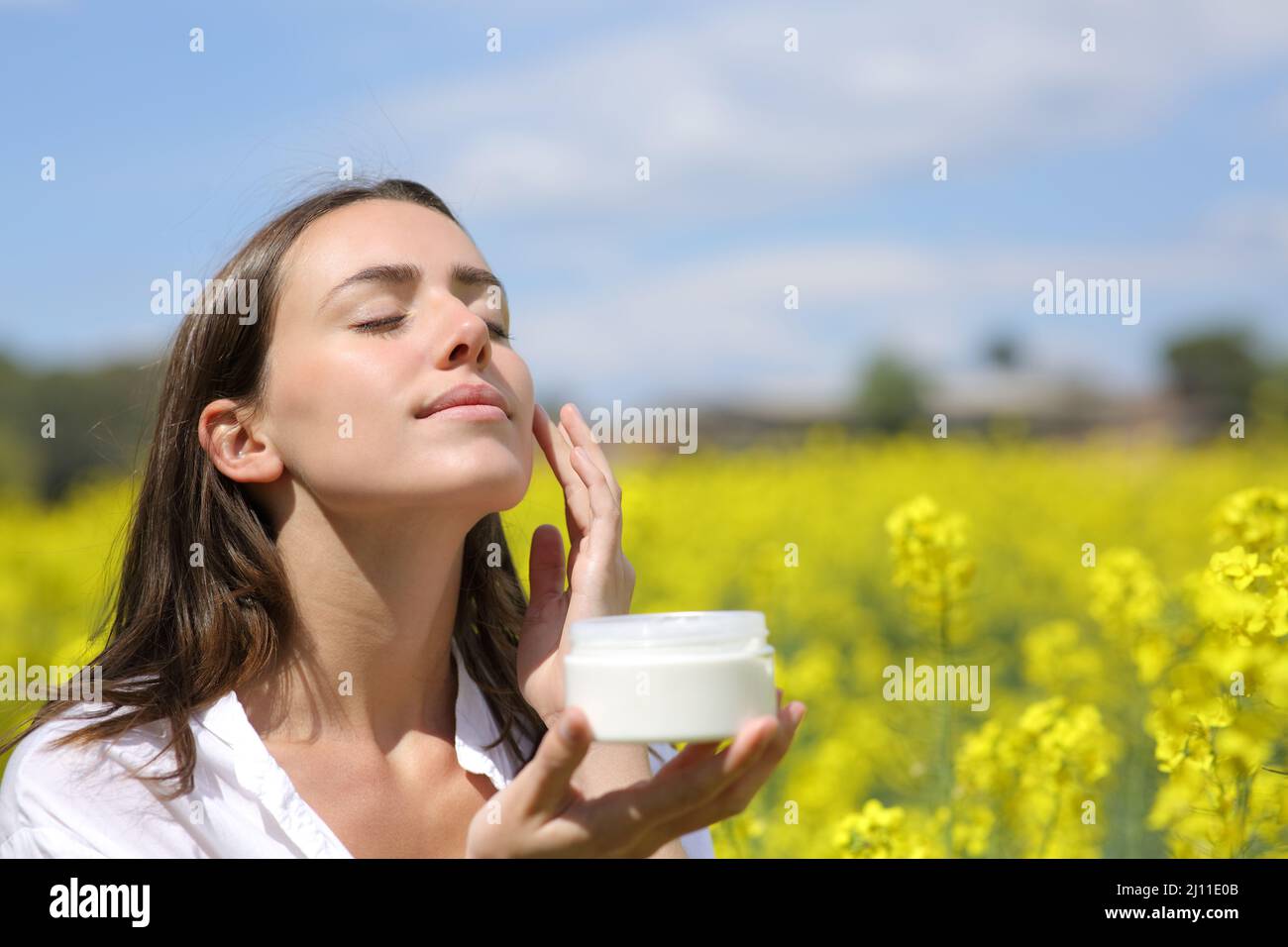 Femme aux yeux fermés appliquant de la crème hydratante sur la joue un jour ensoleillé Banque D'Images
