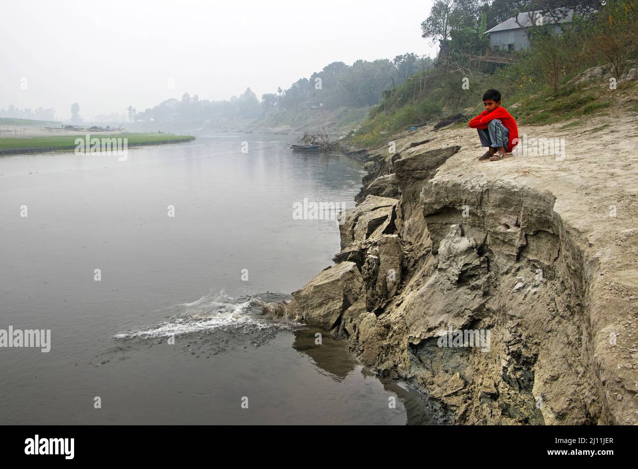 Kamrul, 11 ans, s'assit à côté d'une rive de rivière érodante sur le fleuve Brahmaputra à Manikganj, au Bangladesh, dans des yeux tentares tandis que l'érosion continue de se faire, mettant sa famille au risque de déplacement. La famille de Kamrul vit à côté de la rivière Brahmaputra mais doit partir pour des destinations inconnues après que l'érosion par la rivière a emporté la plupart de leurs terres et de leur maison. Le Bangladesh, avec sa population dense et sa pléthore de rivières, a résisté à plus que sa part de tempêtes tropicales, d'inondations et d'autres catastrophes naturelles. Le changement climatique n'a fait qu'accélérer ces éléments destructeurs. Mais il est aussi confronté à un autre, plus Banque D'Images